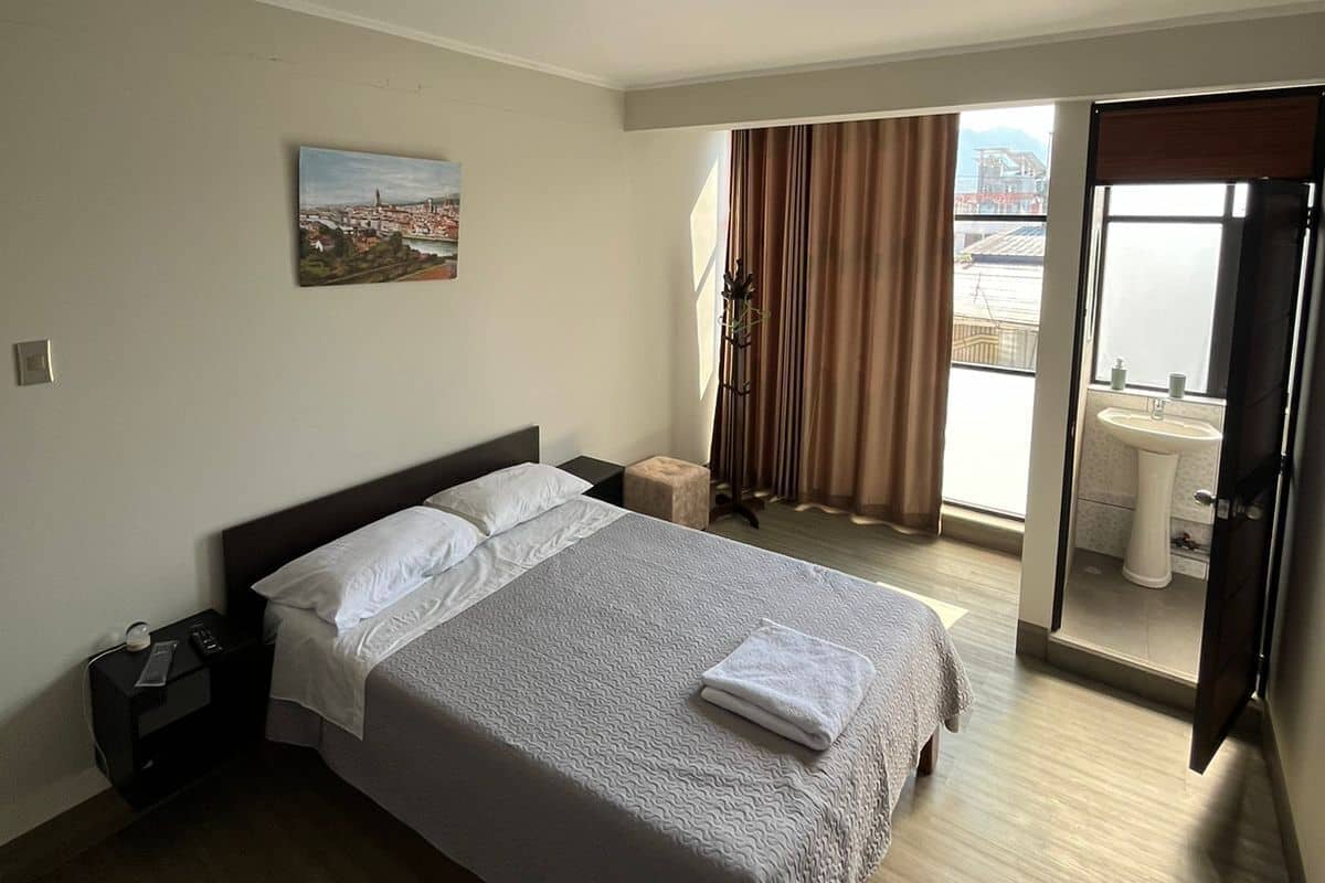 A hotel room with a bed, a nightstand, a painting on the wall, a window with curtains, and a bathroom visible through an open door.