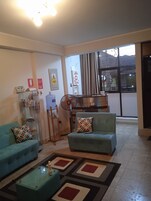 Una sala de estar con dos sofás, una mesa de centro de vidrio y un tapete estampado. Hay letreros en la pared, un ventilador y vistas a través de un ventanal grande.