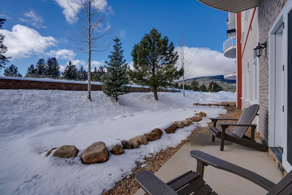 A snow-covered outdoor area with a patio, Adirondack chairs, and a view of a building with a brick wall and a balcony.