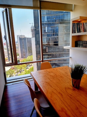 A modern room with a large window, a wooden table, two chairs, a bookshelf, and a potted plant.