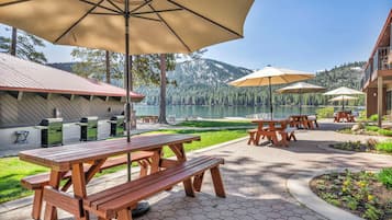 Un área de patio al aire libre con mesas de picnic y sombrillas, con vistas a un lago y montañas al fondo.