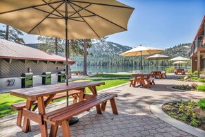 A picnic area with wooden tables and benches, outdoor grills, and umbrellas, overlooking a lake and forested mountains.