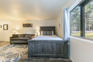 A bedroom with a wooden headboard, a large window with a view of trees, a sofa, and a patterned rug.