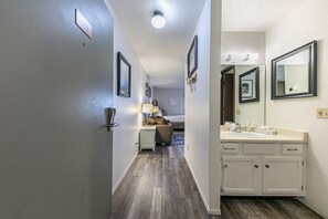 A hotel hallway with a wooden floor, a bed, a nightstand with a lamp, and a bathroom with a sink and mirror.