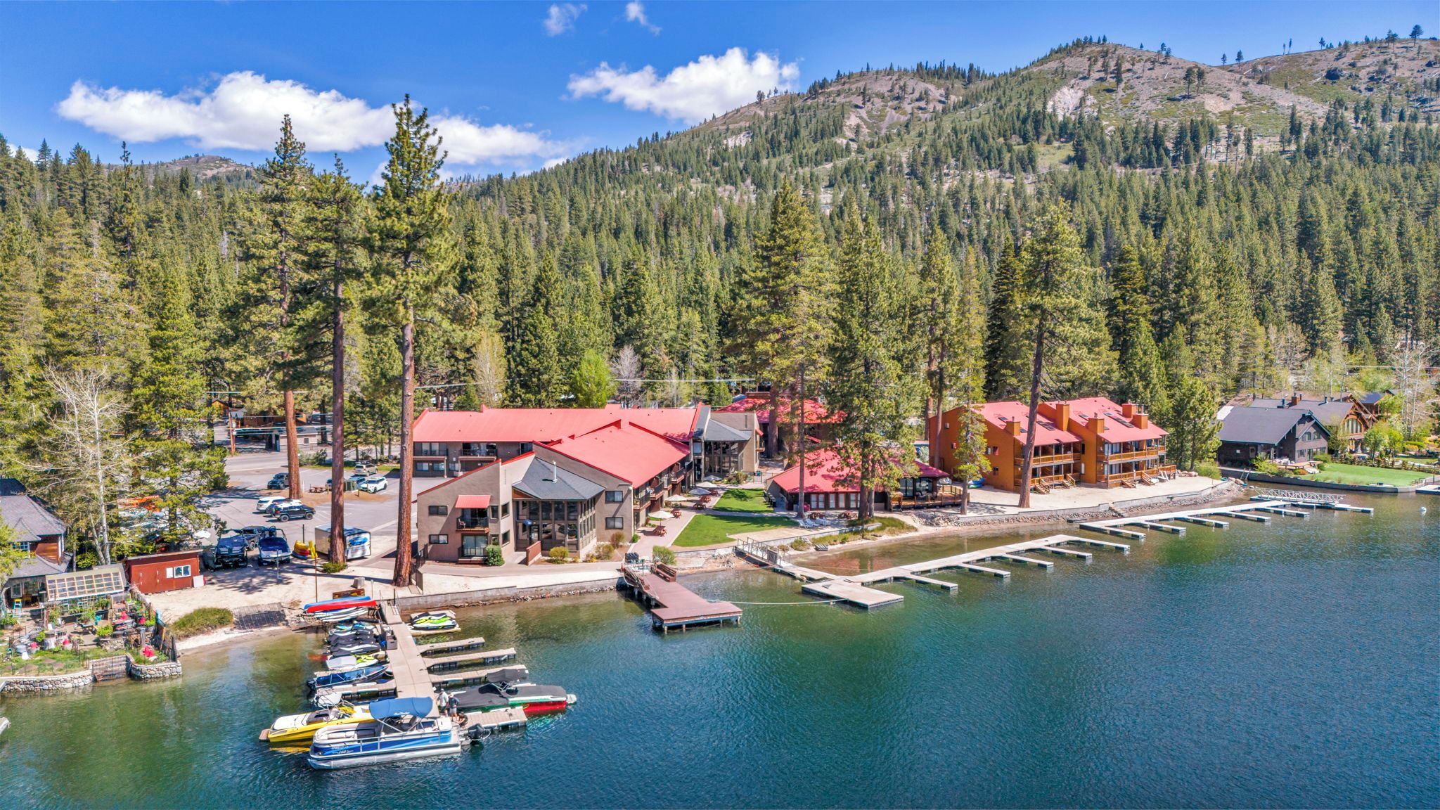 Un lodge junto al lago con varias construcciones, barcos amarrados y una montaña boscosa al fondo.