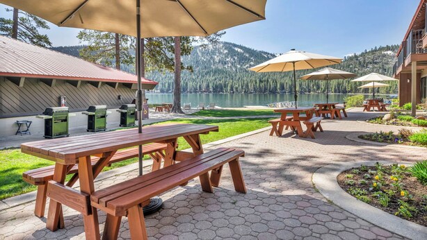 Ein Picknickplatz mit Holztischen und -bänken, Grillstellen und Sonnenschirmen mit Blick auf einen See und bewaldete Berge.