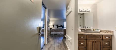 Habitación de hotel con piso de madera, una cama grande con cabecera, un baño con lavamanos y espejo, y un pasillo con una puerta marcada '115'.