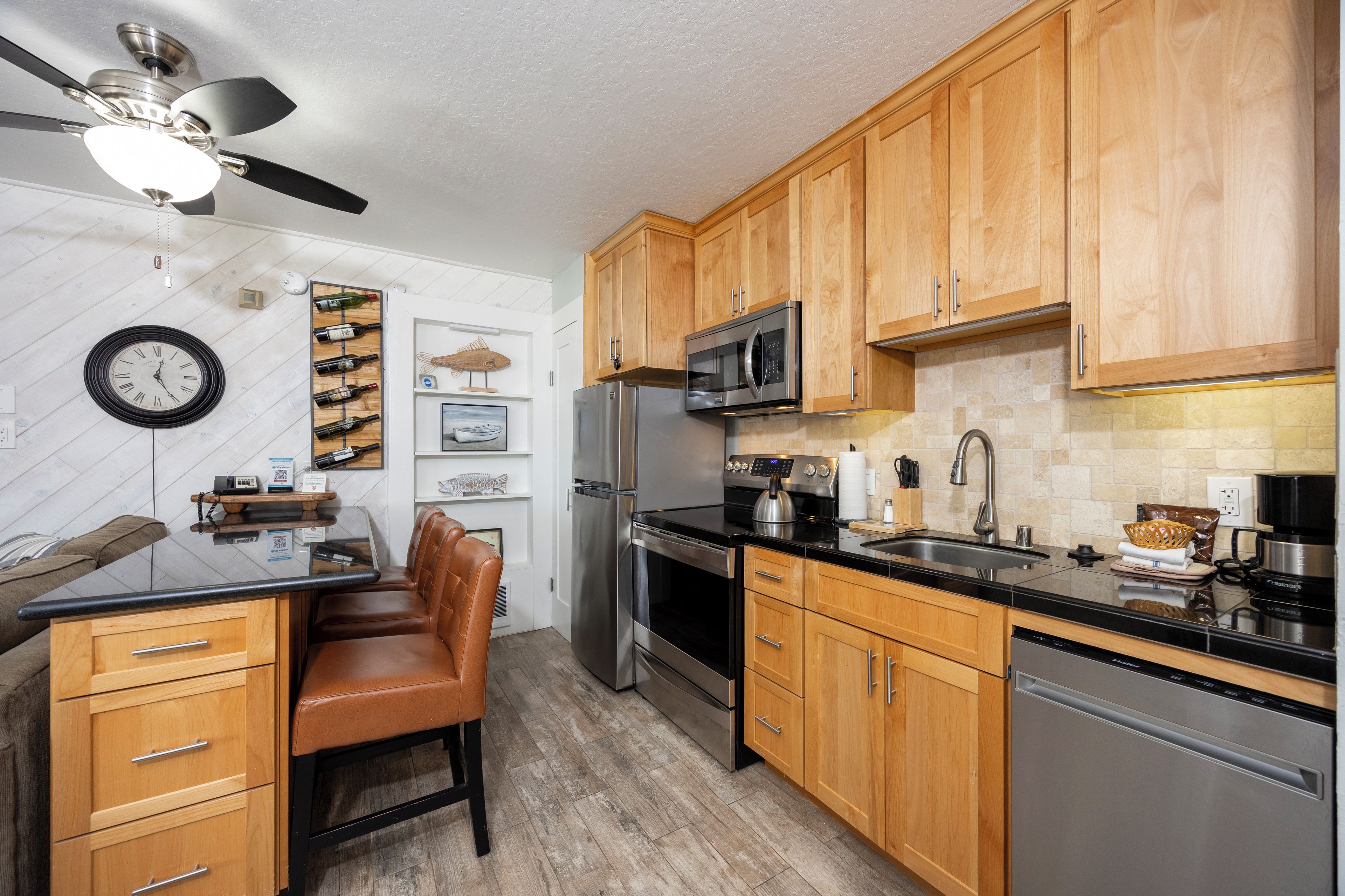 A modern kitchen with wooden cabinets, stainless steel appliances, a black countertop, and a ceiling fan.