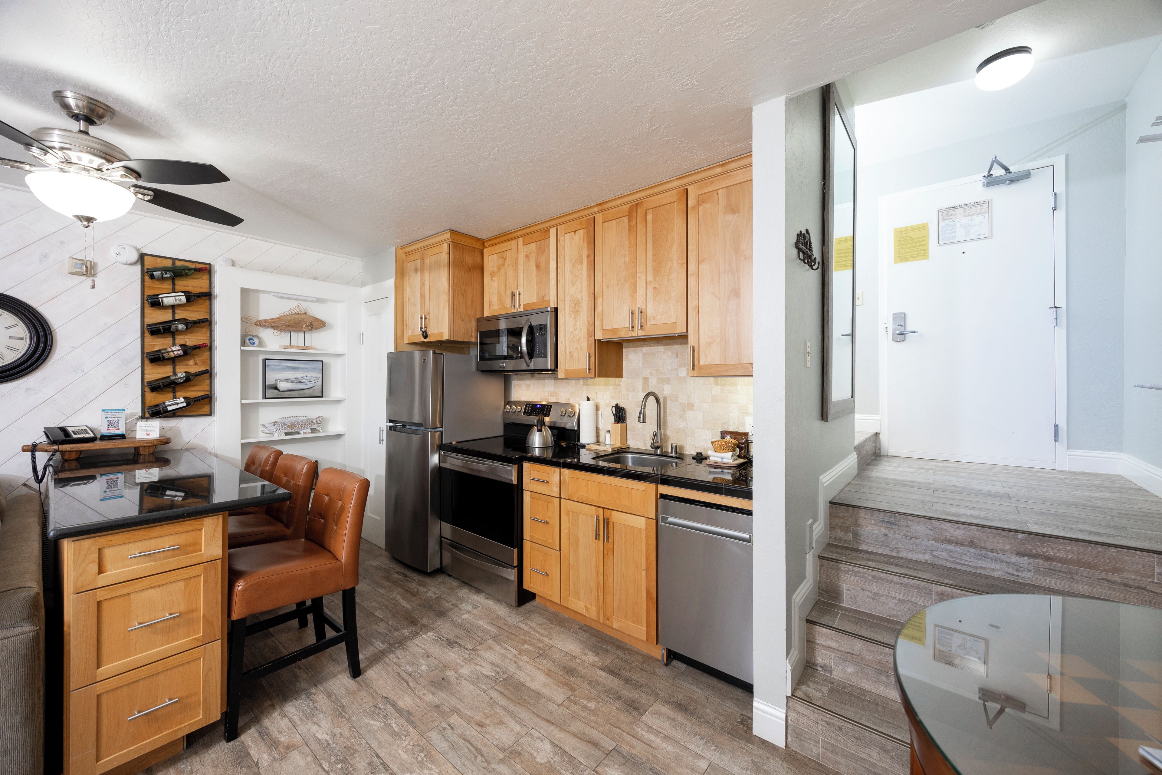 A modern kitchen with wooden cabinets, stainless steel appliances, a dining area with a glass table, and a ceiling fan.