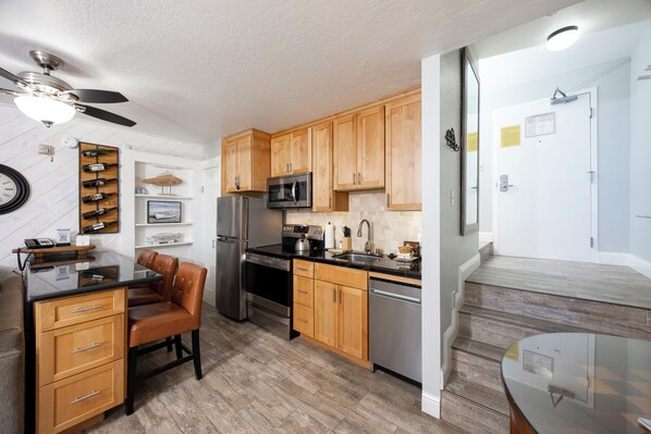 A modern kitchen with wooden cabinets, stainless steel appliances, a dining area with a glass table, and a ceiling fan.