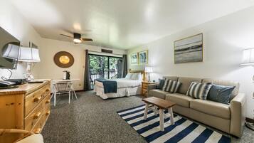 A hotel room with a bed, a sofa, a desk, a chair, a ceiling fan, and a view of the outdoors.