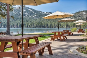 Outdoor wooden picnic tables with umbrellas, a paved walkway, and a lake with mountains in the background. - Cozy Studio w/Kitchenette and Balcony #212 at Donner Lake Village (Truckee)