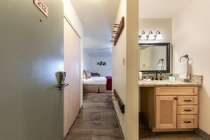 Un couloir d’hôtel menant à une chambre avec un lit, une salle de bain avec un lavabo, un miroir et un porte-serviettes.