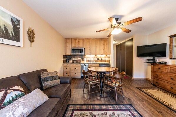 Un salon chaleureux avec un canapé en cuir marron, une table à manger en bois avec des chaises, un ventilateur de plafond et une cuisine dotée de meubles en bois et d’un four à micro-ondes.