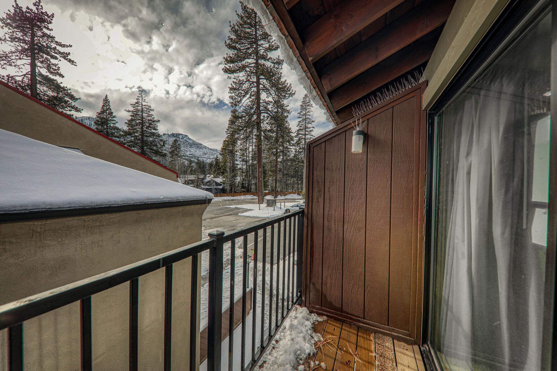 A balcony with a wooden railing, a glass door, and a view of snow-covered ground and trees.