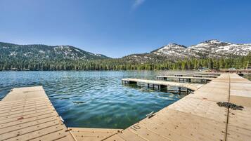 A wooden dock extending into a calm lake with clear blue water, surrounded by forested mountains and snow-capped peaks under a clear sky.
