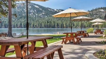 Outdoor wooden picnic tables with umbrellas, a paved walkway, and a lake with mountains in the background.