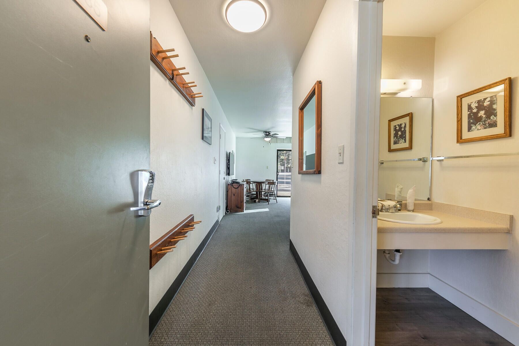 Un couloir d’hôtel avec un sol recouvert de moquette, un porte-manteau, une porte dotée d’un système de verrouillage par carte magnétique, un ventilateur de plafond et une salle de bain équipée d’un lavabo et de tableaux encadrés.