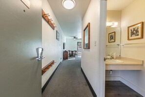 Ein Hotelgang mit Teppichboden, einem Kleiderständer, einer Tür mit Chipkarte, einem Deckenventilator und einem Badezimmer mit Waschbecken und gerahmten Bildern.