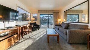 A modern hotel room with a large flat-screen TV, a wooden desk with a laptop, a sofa, and a balcony with a view of snow-covered mountains. - Spacious Condo w/Lake View #136 at Donner Lake Village (Truckee)