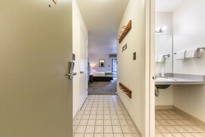 A hotel hallway leading to a bedroom with a bed, a bathroom with a sink and mirror, and a coat rack.