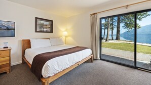 A hotel room with a bed, a bedside table, a lamp, a framed picture, and a large window with a view of trees and a lake. - King Studio w/Lake View #145 at Donner Lake Village (Truckee)
