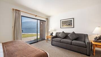 A hotel room with a sofa, a small table with a lamp, a painting on the wall, and a sliding glass door leading to a balcony with a view of the mountains.
