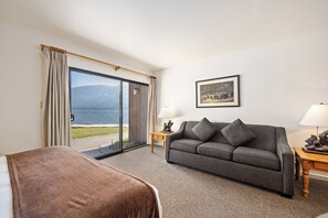 A hotel room with a sofa, a small table with a lamp, a painting on the wall, and a sliding glass door leading to a balcony with a view of the mountains. - King Studio w/Lake View #145 at Donner Lake Village (Truckee)