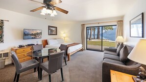 A hotel room with a bed, a sofa, a dining table with chairs, a ceiling fan, and a large window with a view of the outdoors.