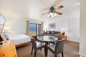 Quarto de hotel com cama, mesa de vidro redonda com cadeiras, ventilador de teto, janela com cortinas, sofá e porta.