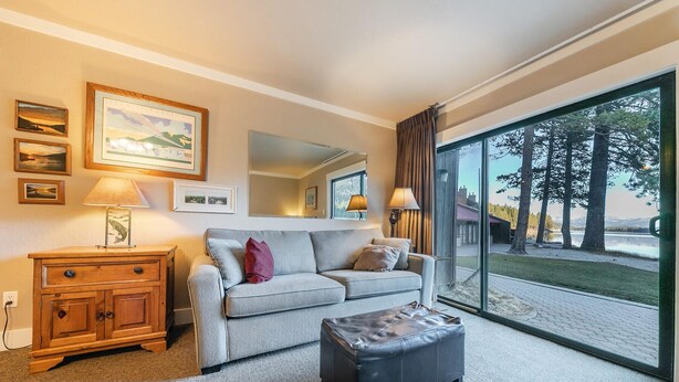 A cozy room with a grey sofa, a wooden cabinet, and a black ottoman. There are framed pictures on the wall, a mirror, and a large window offering a view of trees and a lake.