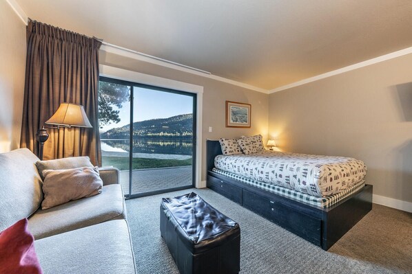 Une chambre d’hôtel avec un canapé, un lit avec une literie à motifs, une petite table et une vue sur un lac, visible à travers une porte coulissante.