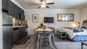 A compact kitchen with modern appliances, a dining area with a table and chairs, a bed with a blue blanket, and a ceiling fan.