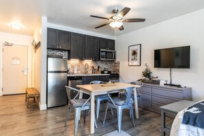 Una cocina compacta con electrodomésticos modernos, un comedor con sillas de metal, un ventilador de techo y un televisor de pantalla plana.