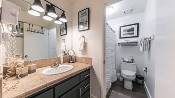 A modern bathroom with a beige countertop, a white sink, and a large mirror. There are three pendant lights above the mirror, and a framed picture of Lake Tahoe on the wall.
