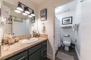 Une salle de bain moderne avec un comptoir beige, un lavabo blanc et un grand miroir. Trois lampes suspendues sont fixées au-dessus du miroir, et un tableau encadré représentant le lac Tahoe est accroché au mur.