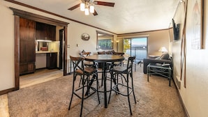 A spacious living area with a dining table, chairs, a sofa, and a ceiling fan. There is a kitchen area with cabinets and a refrigerator. A large window provides a view of a lake and mountains.