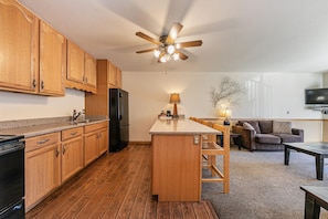 A modern kitchen with wooden cabinets, a black refrigerator, and a central island. There is a ceiling fan, a dining area with a table and chairs, and a living room with a sofa and a TV. - Spacious 1BR Condo w/Partial Lake View #222 at Donner Lake Village (Truckee)