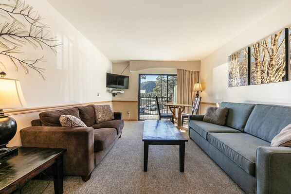 Una sala de estar moderna con un sofá, una mesa de centro y un televisor. Hay un balcón con vistas al exterior.
