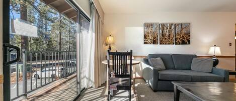 Ein modernes Wohnzimmer mit einem grauen Sofa, einem Couchtisch und einem Sessel. Ein großes Fenster bietet Blick auf Bäume und einen Parkplatz. An der Wand hängt ein Baumgemälde.