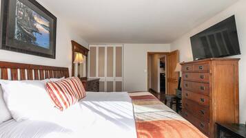 A hotel room with a bed, a wooden dresser, a desk, a chair, a television, and a wardrobe.
