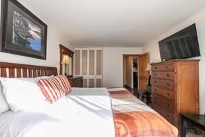 A hotel room with a bed, a wooden dresser, a desk, a chair, a television, and a wardrobe.