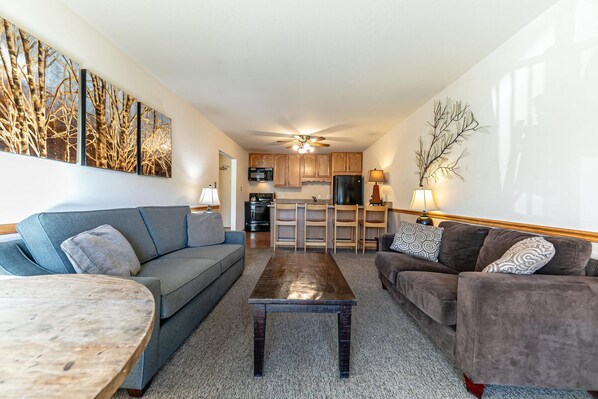 A living room with a grey sofa, a brown sofa, a coffee table, and a dining table. There is a kitchen in the background with wooden cabinets and a black refrigerator.