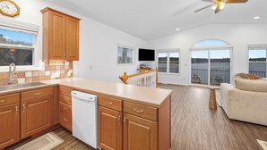 A spacious kitchen with wooden cabinets, a dishwasher, and a large island. There is a ceiling fan, a TV mounted on the wall, and a balcony with outdoor furniture.