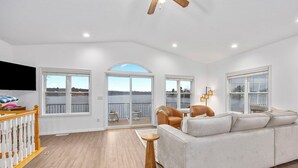 A spacious living room with a large sofa, a television, and a balcony with a view of the water.
