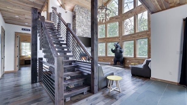 A spacious interior with a wooden staircase, a stone fireplace, and large windows offering natural light.