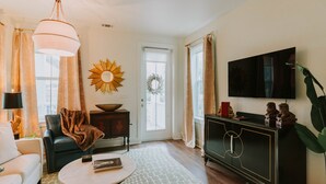 A living room with a sofa, a coffee table, a TV, a black cabinet, and a large window with curtains. - Mermaid's Muse - Includes GOLF CART! Close to beach & downtown Ocean Springs! (Ocean Springs)