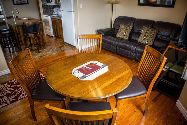 Un comedor con una mesa redonda de madera rodeada de sillas de madera, una cocina al fondo y un sofá de cuero con cojines estampados.