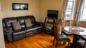 Una sala de estar con un sofá de cuero, un televisor sobre un mueble bajo, una mesa de comedor con sillas y una ventana con cortinas.