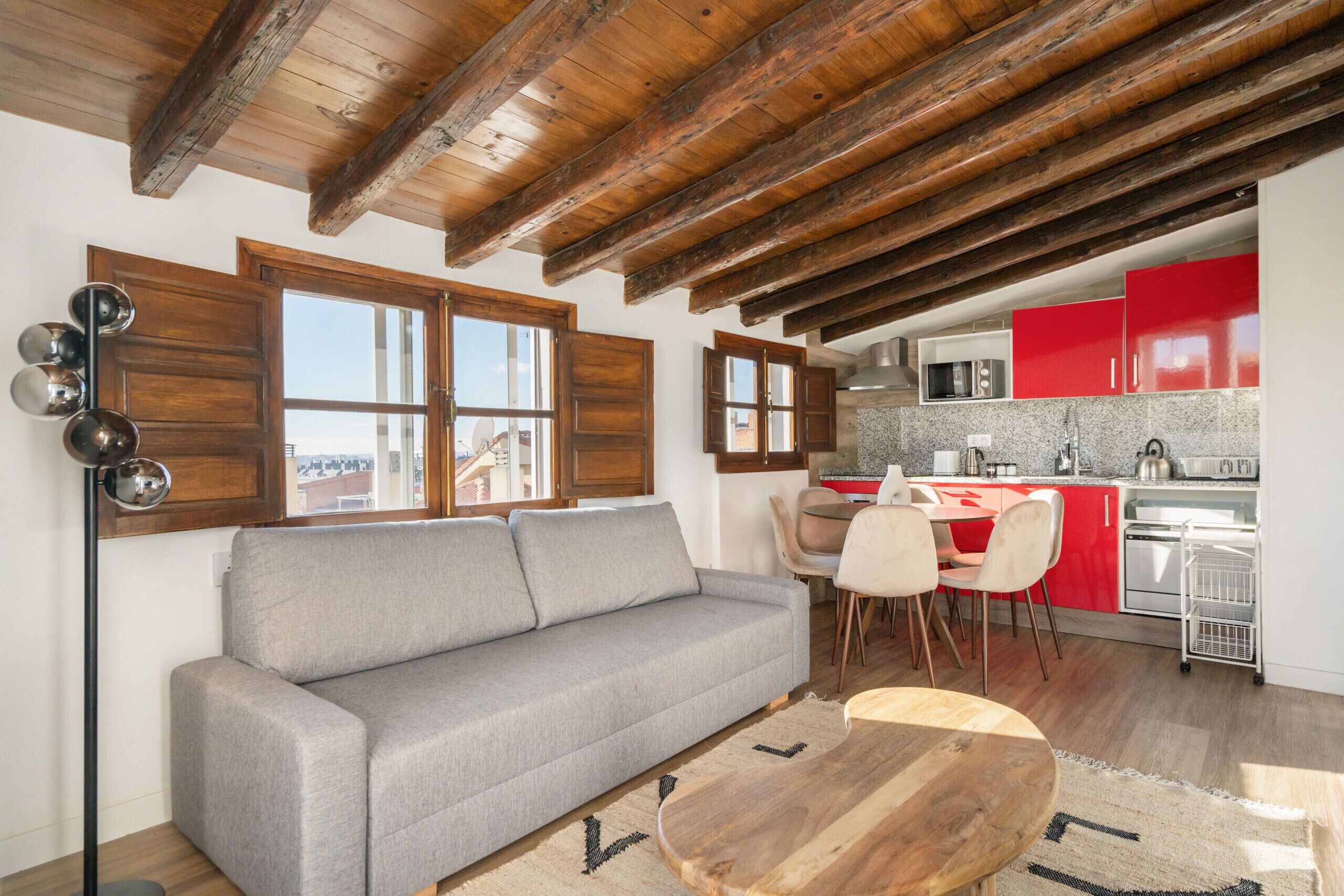 A cozy living room with a grey sofa, wooden ceiling beams, and a dining area with red cabinets and a white kitchen.